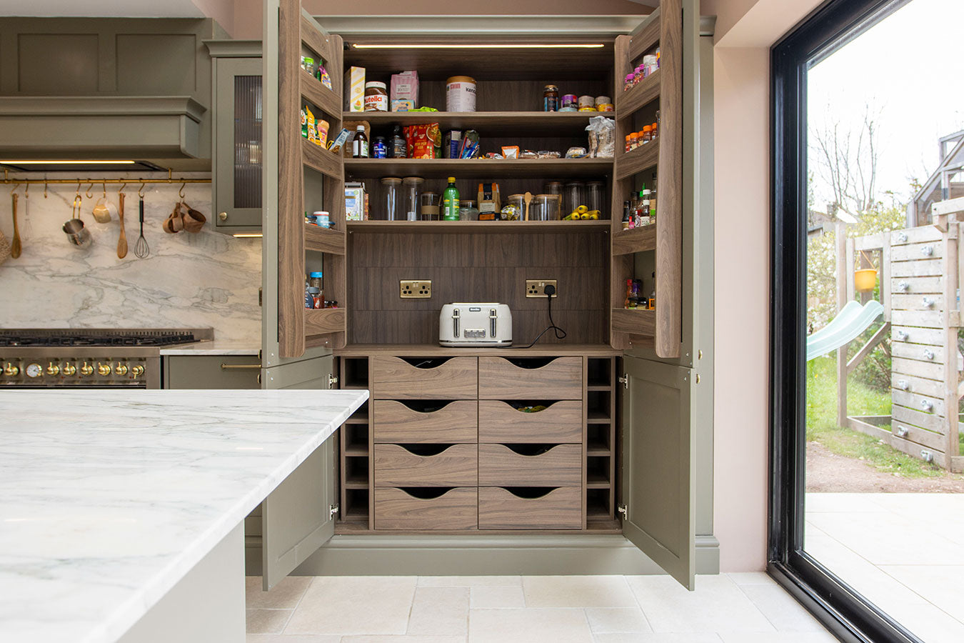 Open kitchen pantry with shelves and drawers filled with food items, located next to a window.